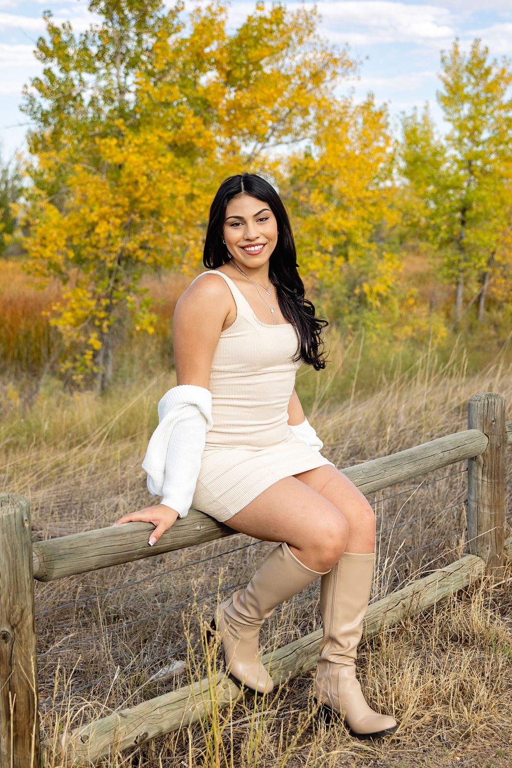 Person in cream dress and boots sits on wooden fence with autumn foliage in background.