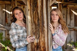 Two young women in plaid shirts pose against a rustic wooden pole in a barn setting.