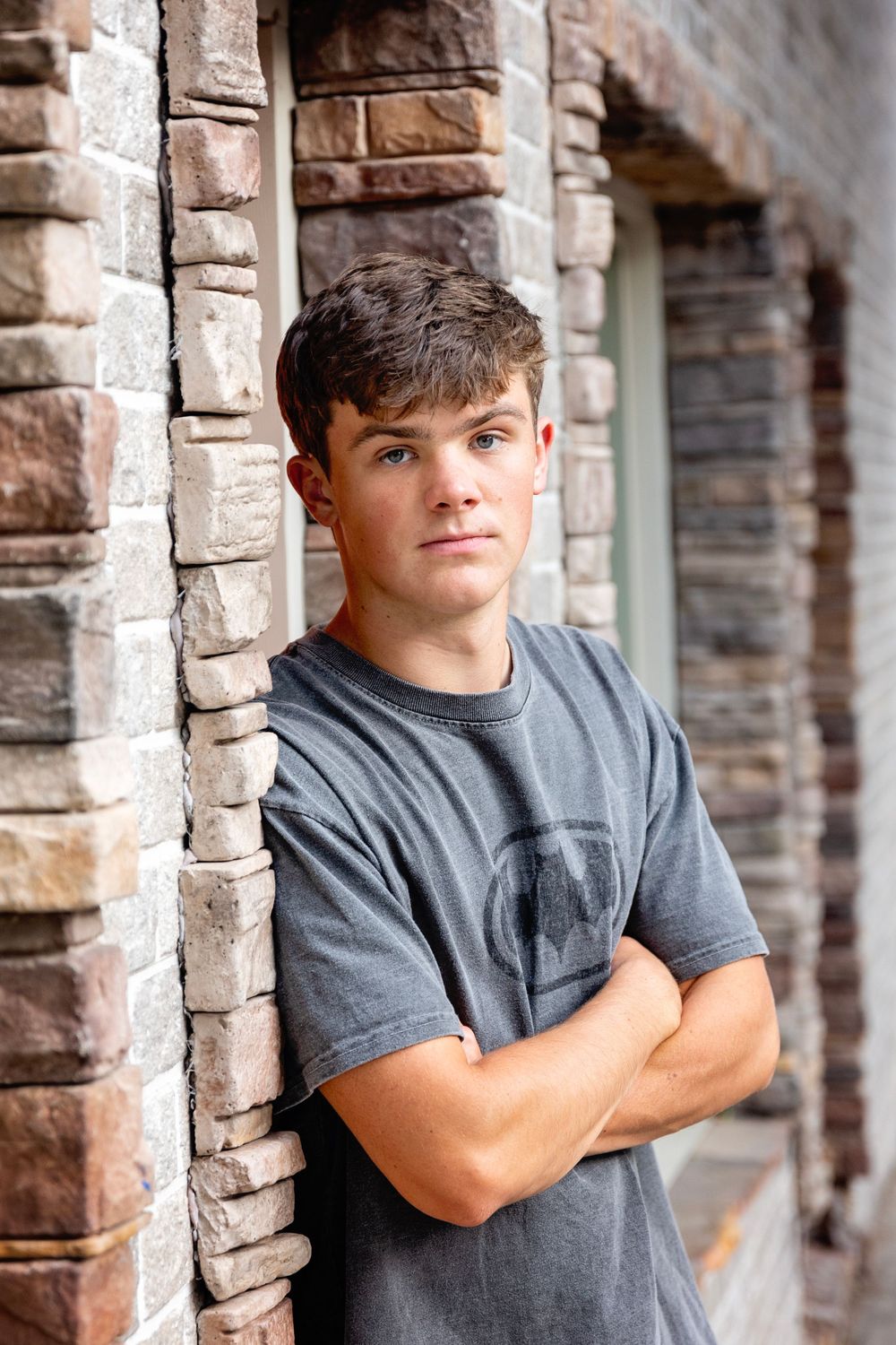 Person wearing a gray t-shirt poses against brick wall columns with crossed arms in outdoor portrait setting.