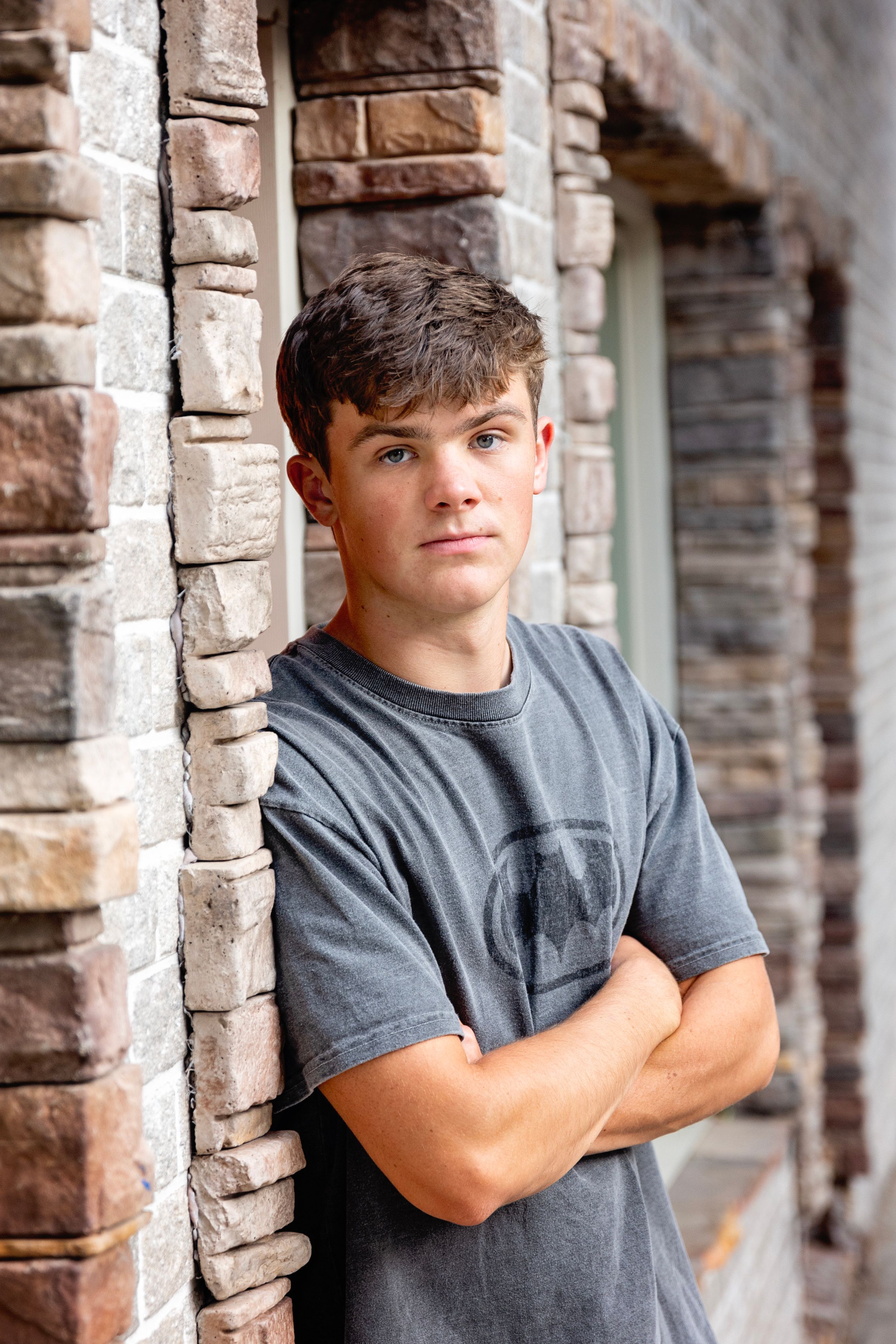 Person wearing a gray t-shirt poses against brick wall columns with crossed arms in outdoor portrait setting.