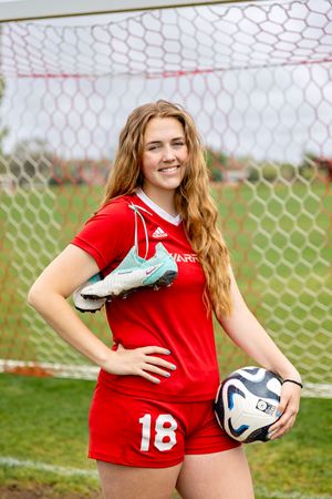 Soccer player in red Adidas uniform holds soccer ball while posing in front of goal net on grass field.