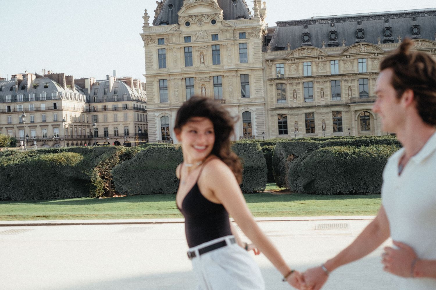 Two people run playfully in front of an ornate historic French building with classical architecture and manicured hedges.