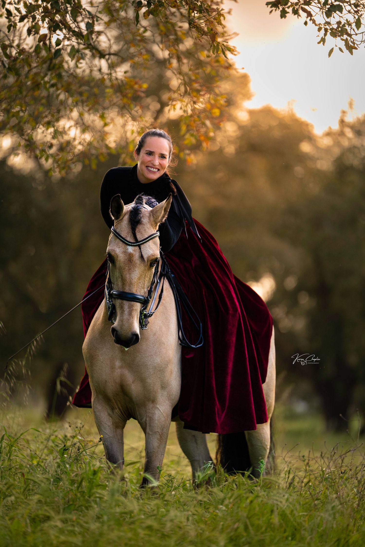 Smiling woman in a deep red velvet skirt riding a buckskin horse through a sunlit field, equine portrait by Karen Chaplin Photography