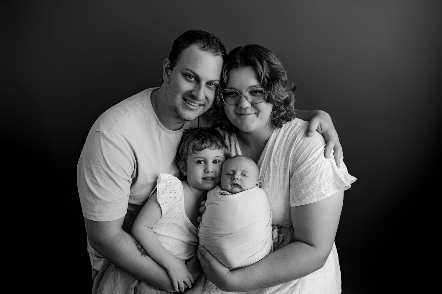 Black and white studio portrait of a couple in white shirts embracing with two children in soft focus against dark background.
