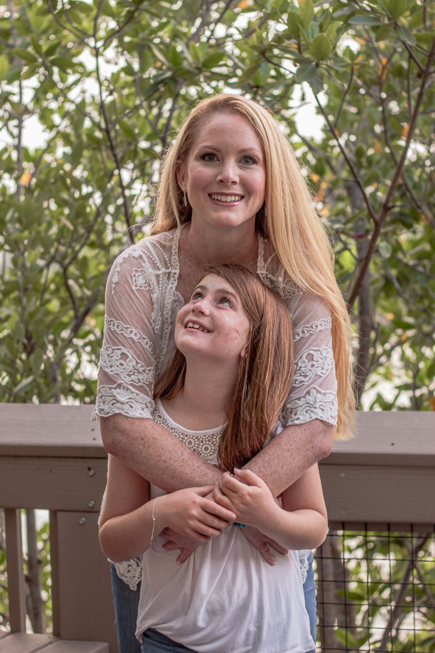 A mother and daughter share a tender embrace on a wooden deck surrounded by green foliage.