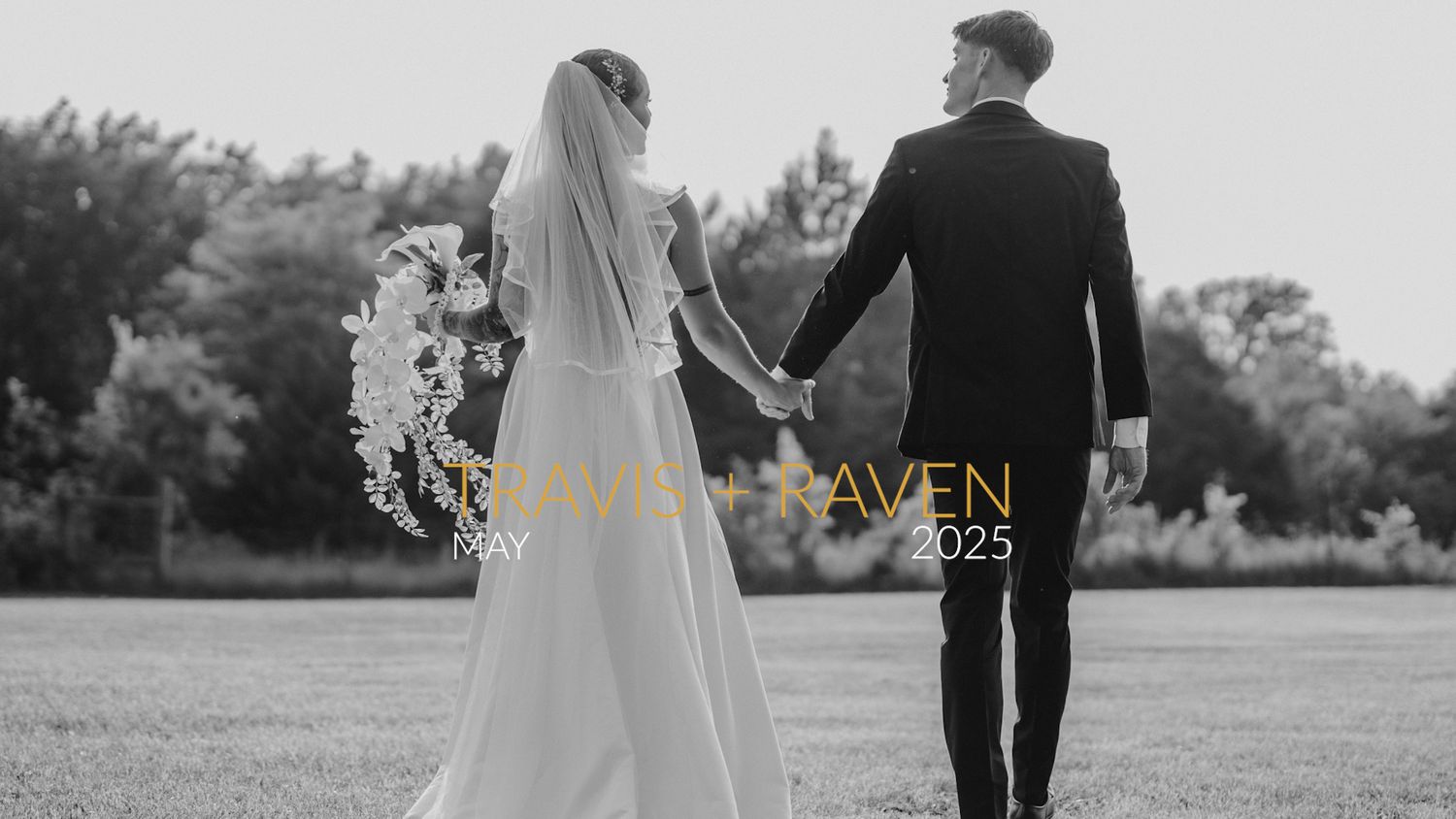 A black and white wedding portrait captures a couple walking hand in hand across an open field.