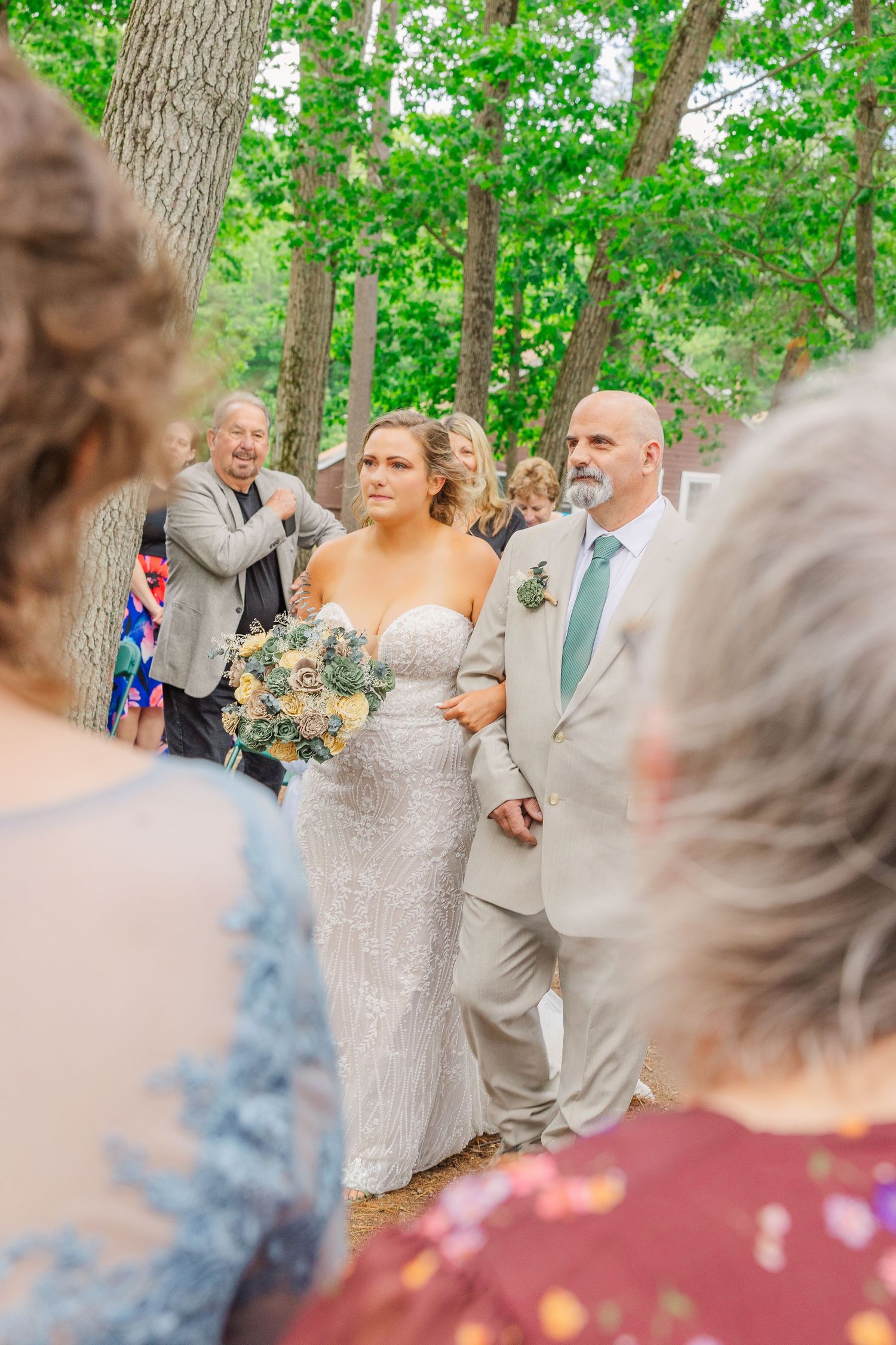 Groomsmen in matching light gray suits with yellow boutonnieres stand together during an outdoor woodland wedding ceremony.