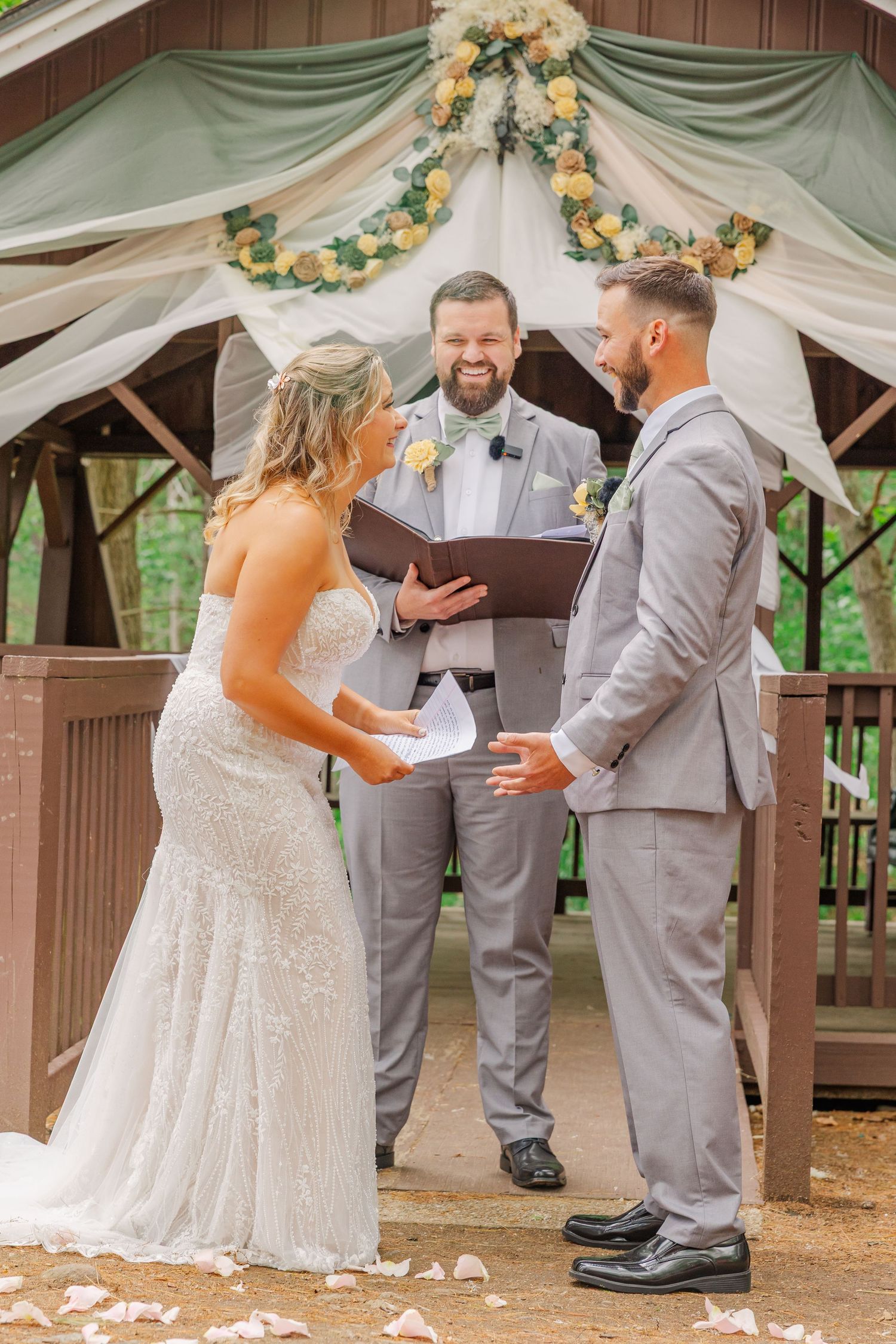 A wedding ceremony takes place in a decorated gazebo with white drapery and yellow floral arrangements overhead.
