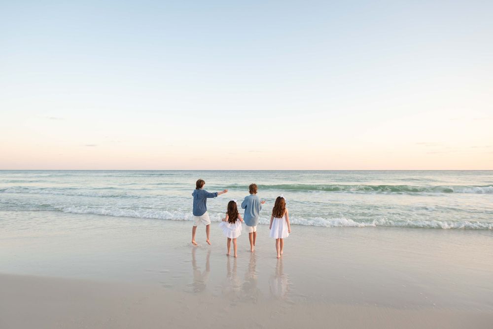 Three people stand together at the edge of the ocean waves on a sandy beach during sunset.