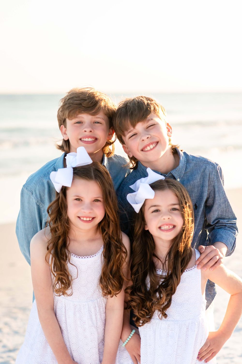 Four children in coordinated blue and white outfits pose together smiling on a sunny beach.