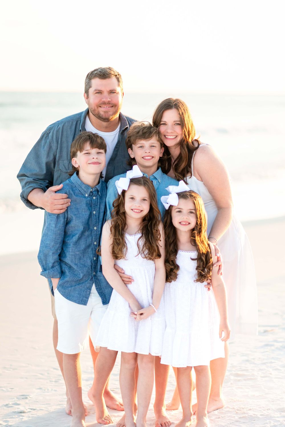 Family dressed in coordinating blue and white outfits poses together on sunny beach at sunset.