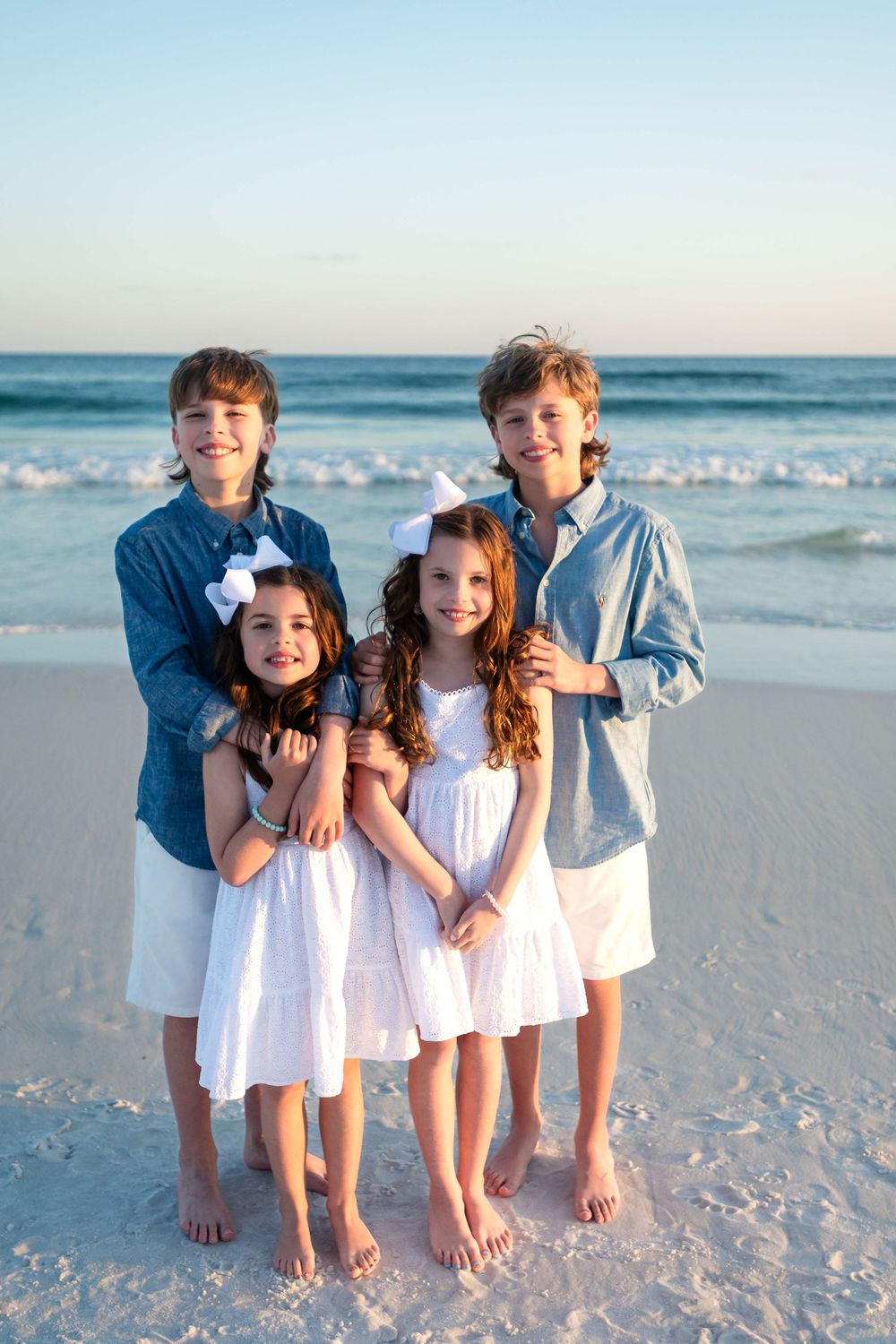 Children in white clothes playing and posing together on a sunny beach with ocean waves in the background.