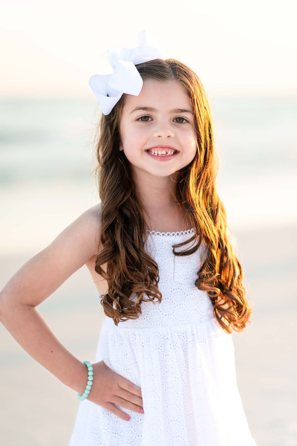 Long-haired child in white eyelet dress wearing a white bow and turquoise bracelet poses at beach during golden hour.