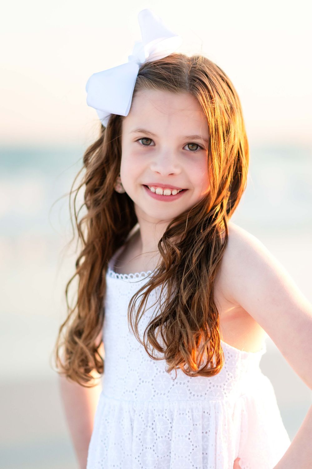 Young child in white eyelet dress and blue hair bow poses on beach during golden hour lighting.