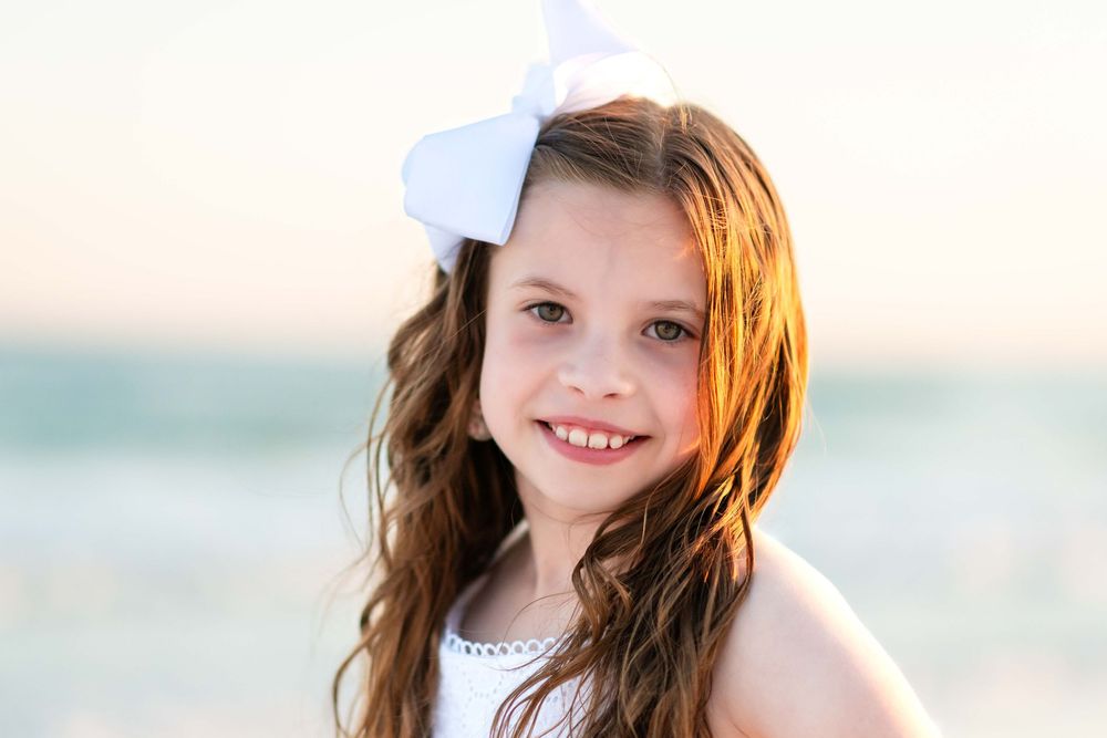 Young person wearing a white hair bow smiles in warm sunset light at the beach.