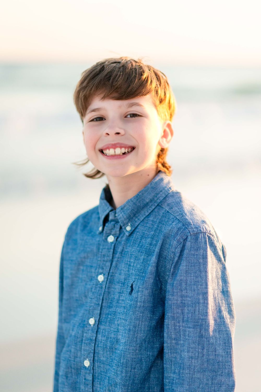 Young person wearing a chambray button-down shirt smiles warmly against a soft, blurred background.