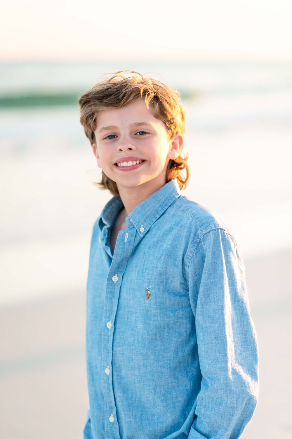 Child in light blue button-down shirt smiling on beach with soft sunset lighting in background.