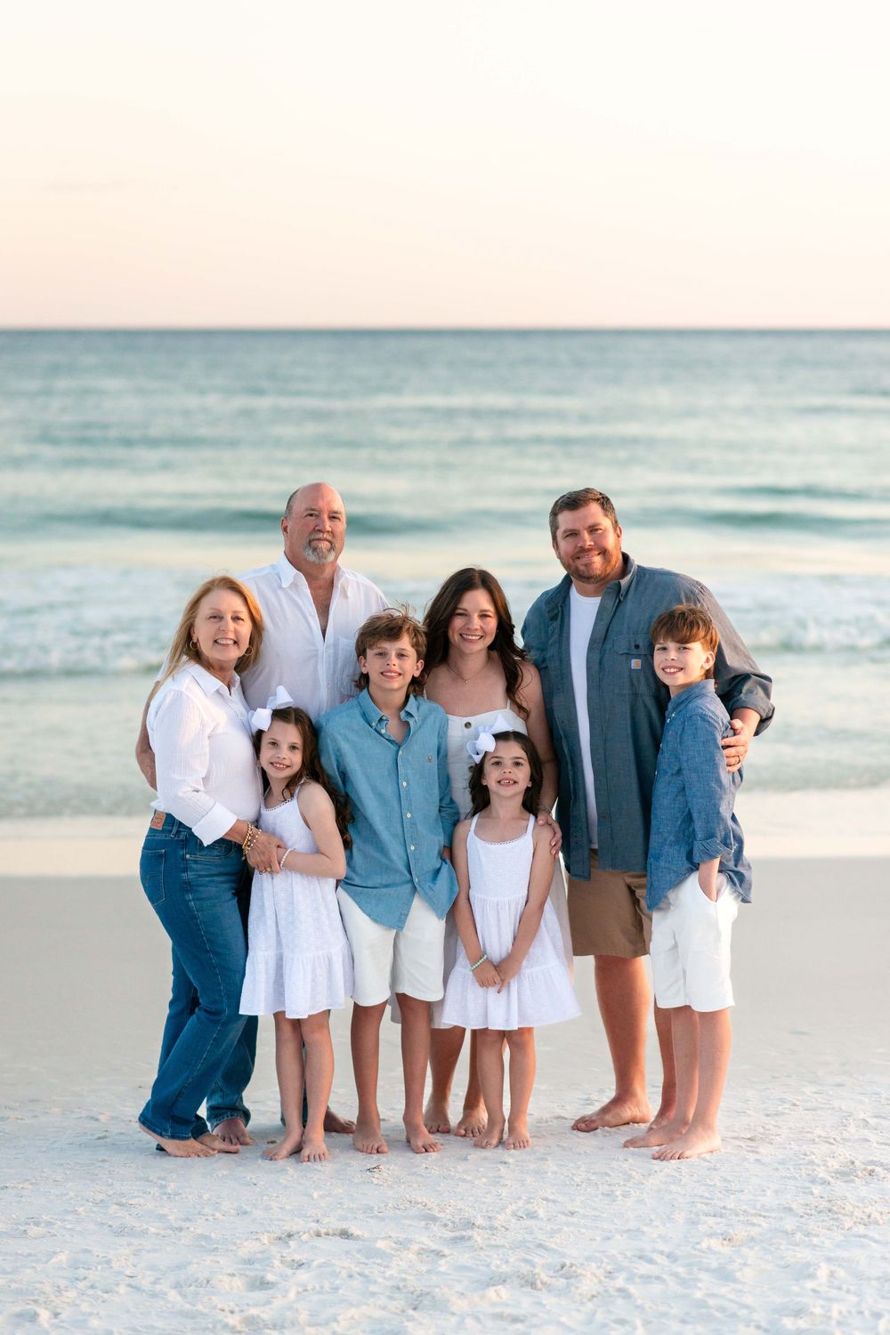 A group of people wearing coordinated blue and white outfits pose together on a beach at sunset.