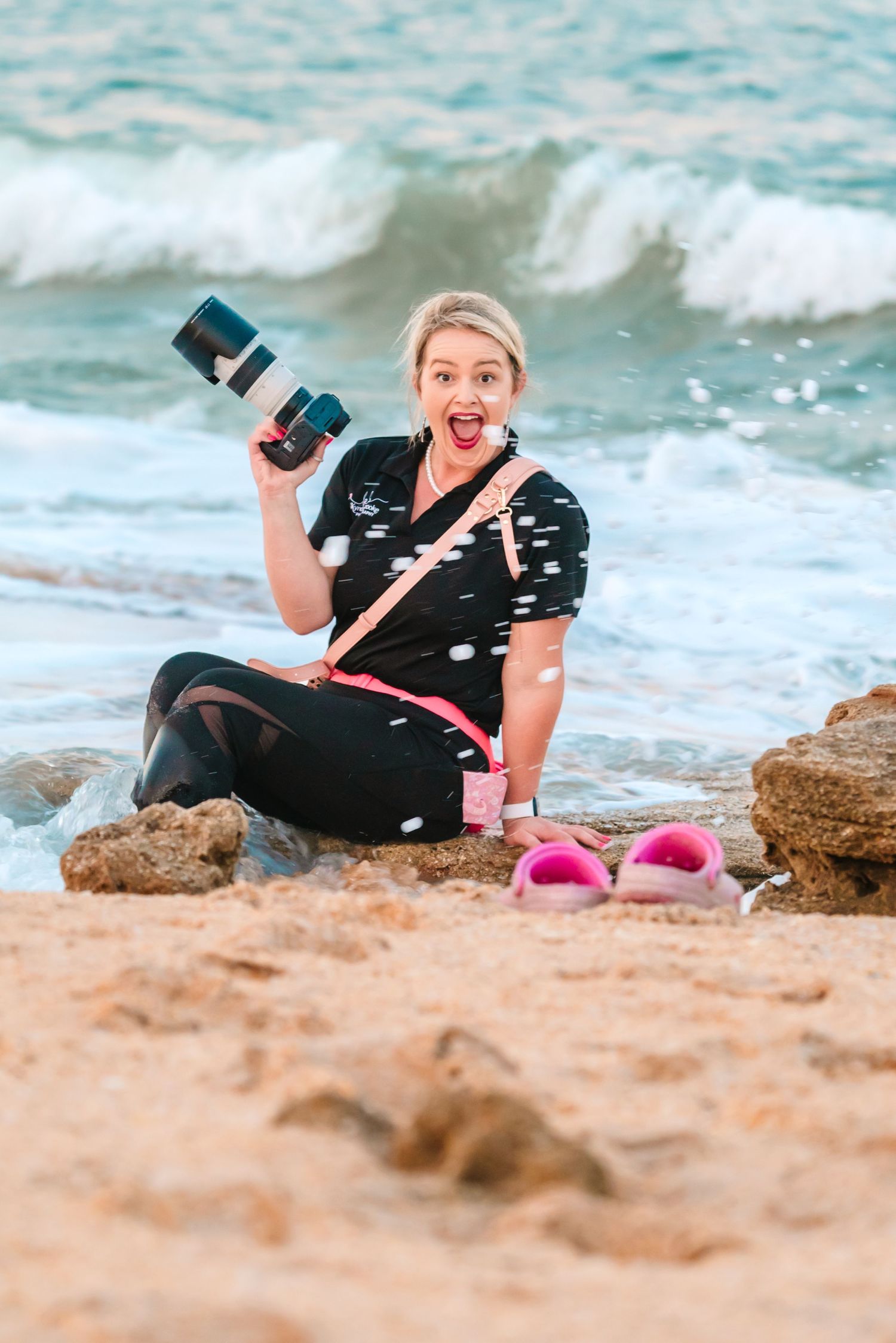 Photographer sits on beach rocks with camera gear and pink sandals while ocean waves crash in background.