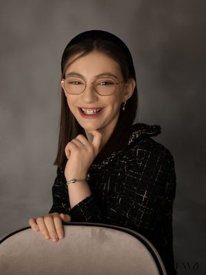 A portrait of someone in glasses and a black textured jacket smiling warmly while posing against a gray background.