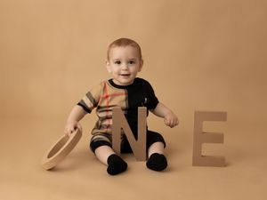 Studio portrait with decorative 'ONE' text props and black onesie against a warm tan background.