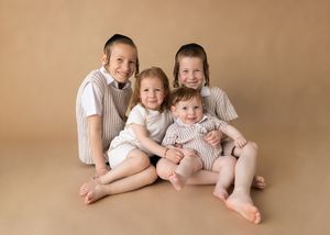 Four young siblings wearing matching light colored outfits sit closely together on a tan backdrop in a natural studio pose.