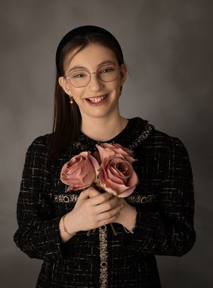 A person wearing glasses and a black textured sweater holds two pink roses while smiling against a gray backdrop.