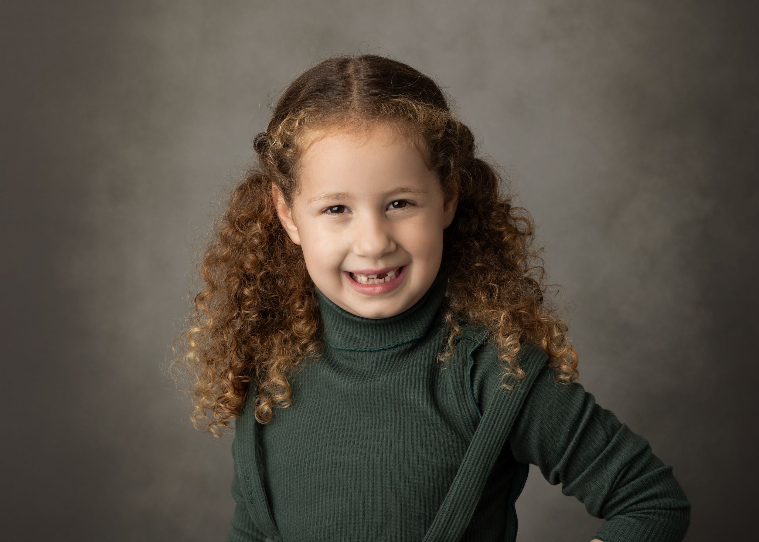 Child with curly hair wearing green turtleneck sweater against grey studio background.
