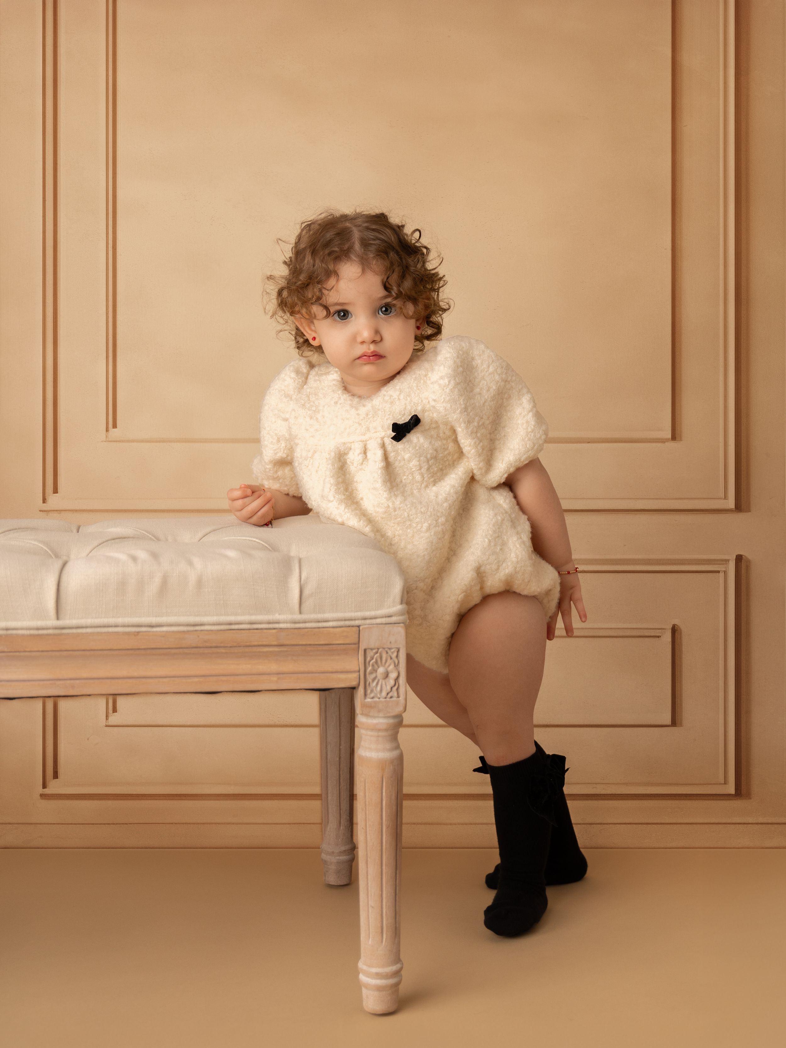 Small child wearing cozy cream sweater outfit poses on vintage white bench against beige paneled wall in studio portrait.