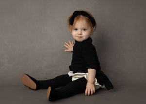 Baby in black outfit with bow sitting on grey studio floor.