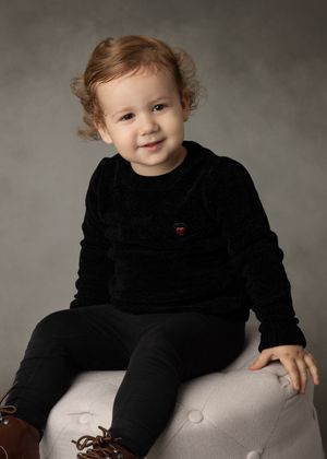 Young child in black velvet outfit sitting on grey ottoman against neutral backdrop.