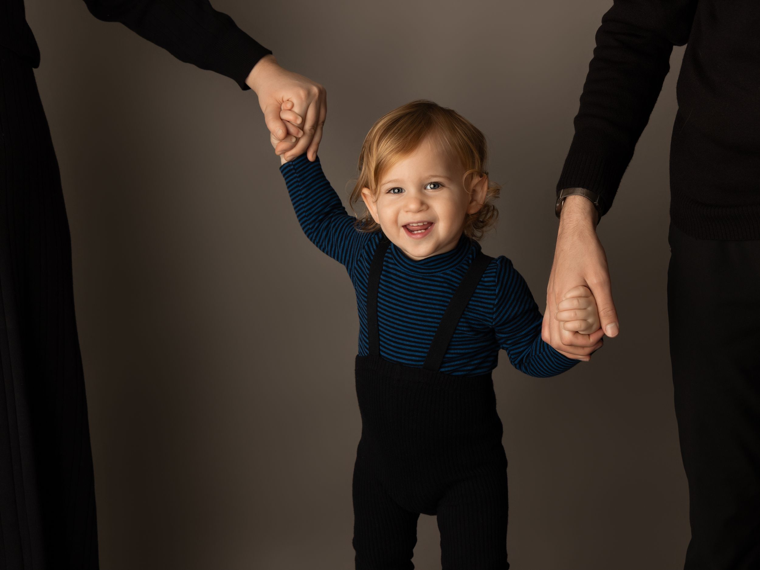 Joyful toddler in a navy and black outfit holds hands with adults while standing against a gray background.