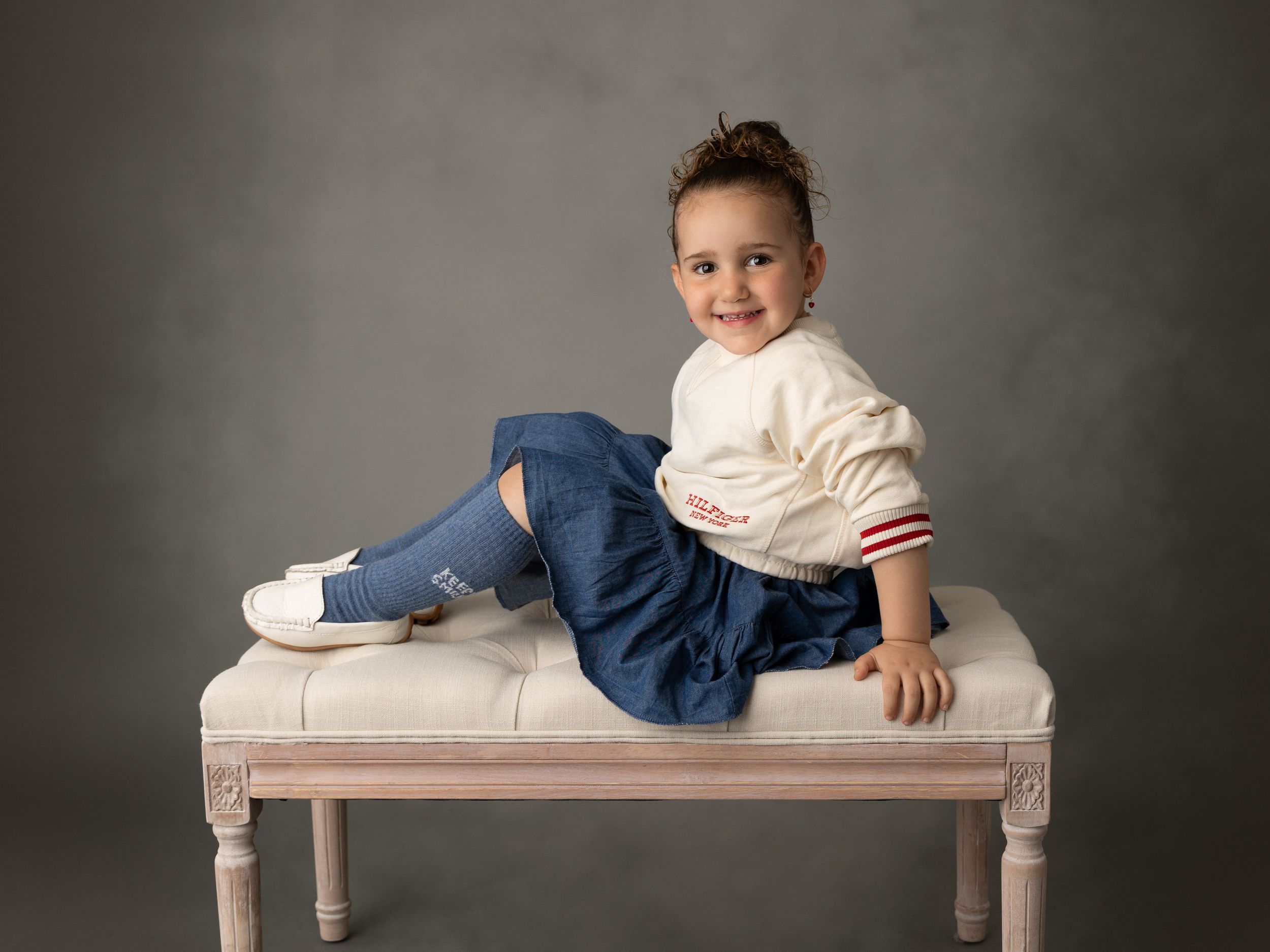 Young child in casual attire relaxing on vintage-style bench against grey backdrop.