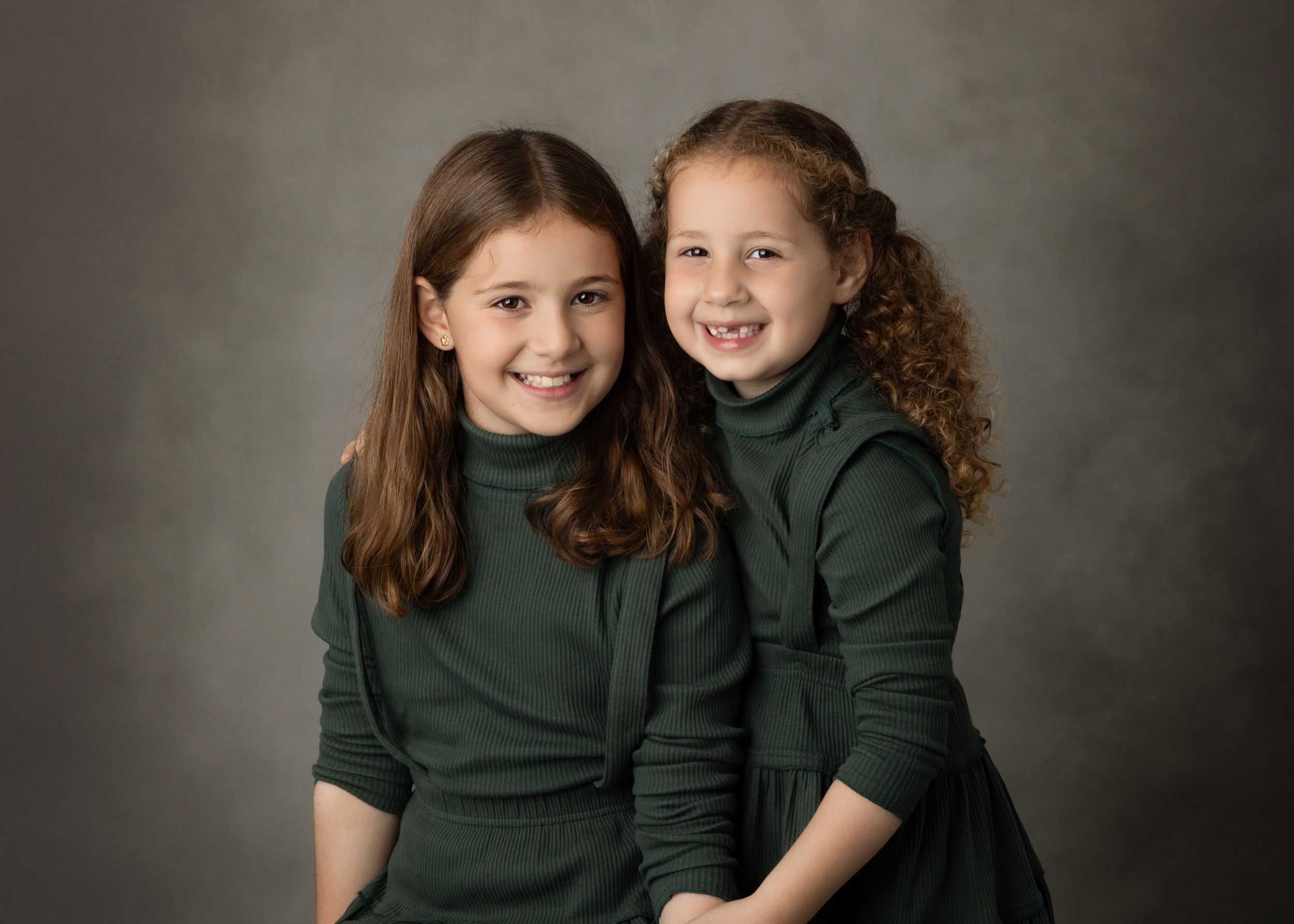 Two people in matching dark green tops share a warm embrace against a neutral studio backdrop.