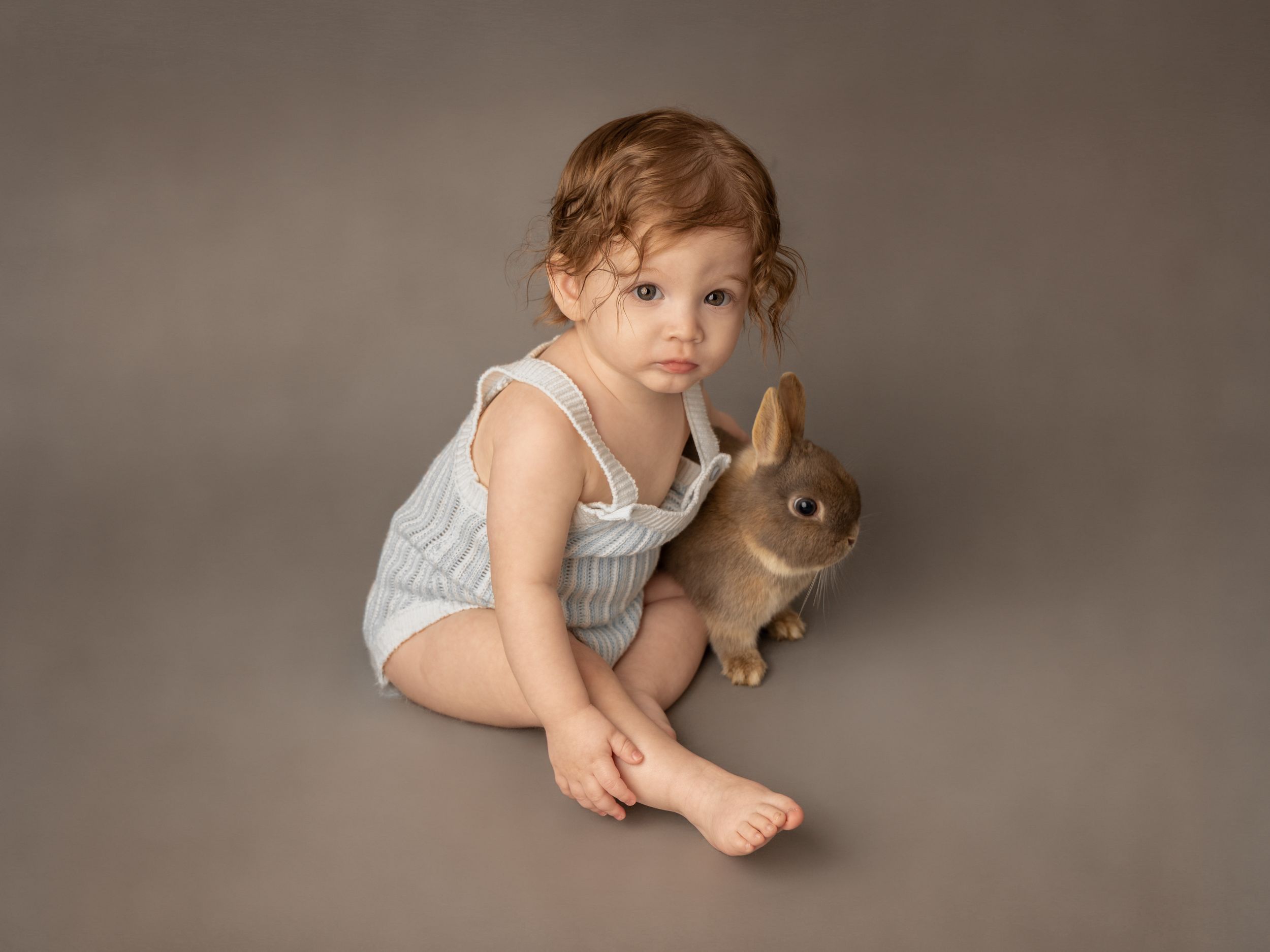 A toddler in a gray romper sits beside a brown rabbit against a neutral studio background.