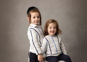 Two young children in matching white hoodies with navy stripes smile while posing for a portrait against a gray backdrop.