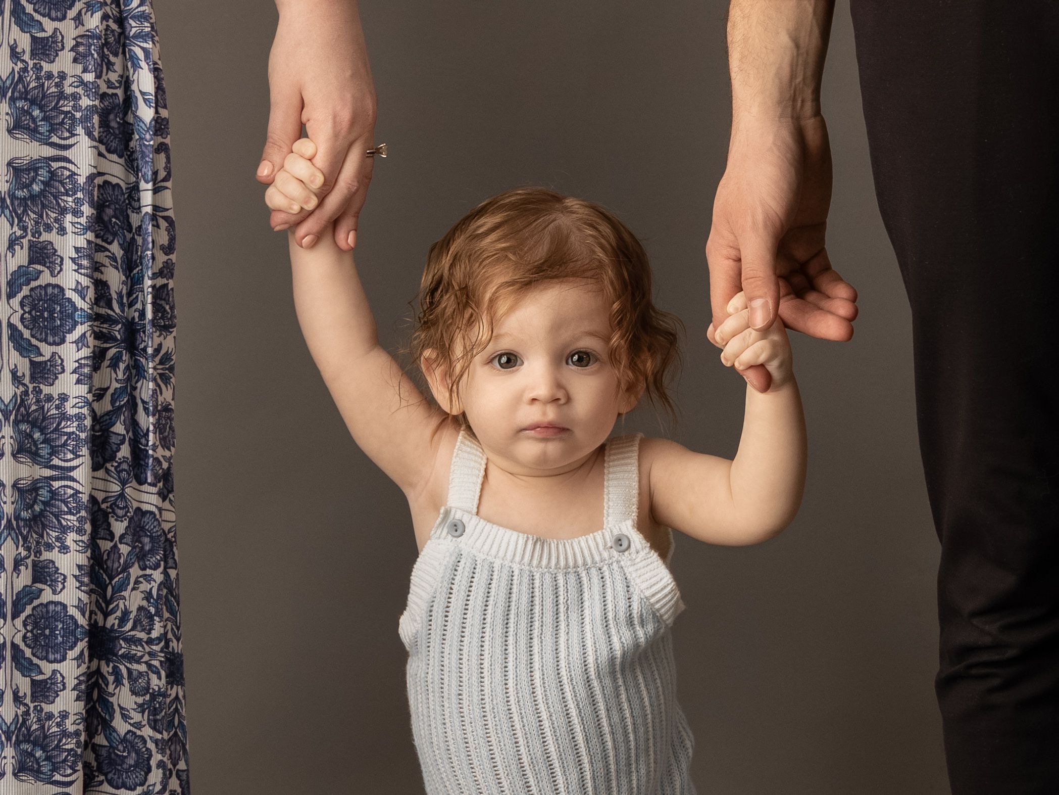 Toddler in gray romper holds hands with two adults during a professional studio portrait session.