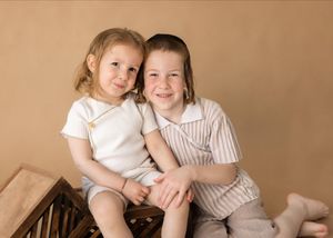 Two young children in light-colored clothing sit together against a brown studio backdrop.