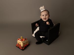 A child wearing formal attire and crown sits in a black chair next to a decorated holiday gift box.