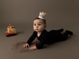 Small child in black outfit and paper crown lying on stomach on neutral backdrop with toy car nearby.