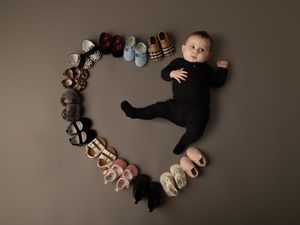 Baby in black outfit lies inside heart shape made of arranged baby shoes on brown background.