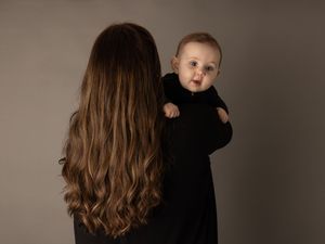 A baby in a black outfit reaches out while being held by someone with long wavy brown hair against a neutral backdrop.