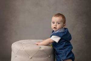 Small child in blue outfit stands beside beige upholstered ottoman against gray studio background.