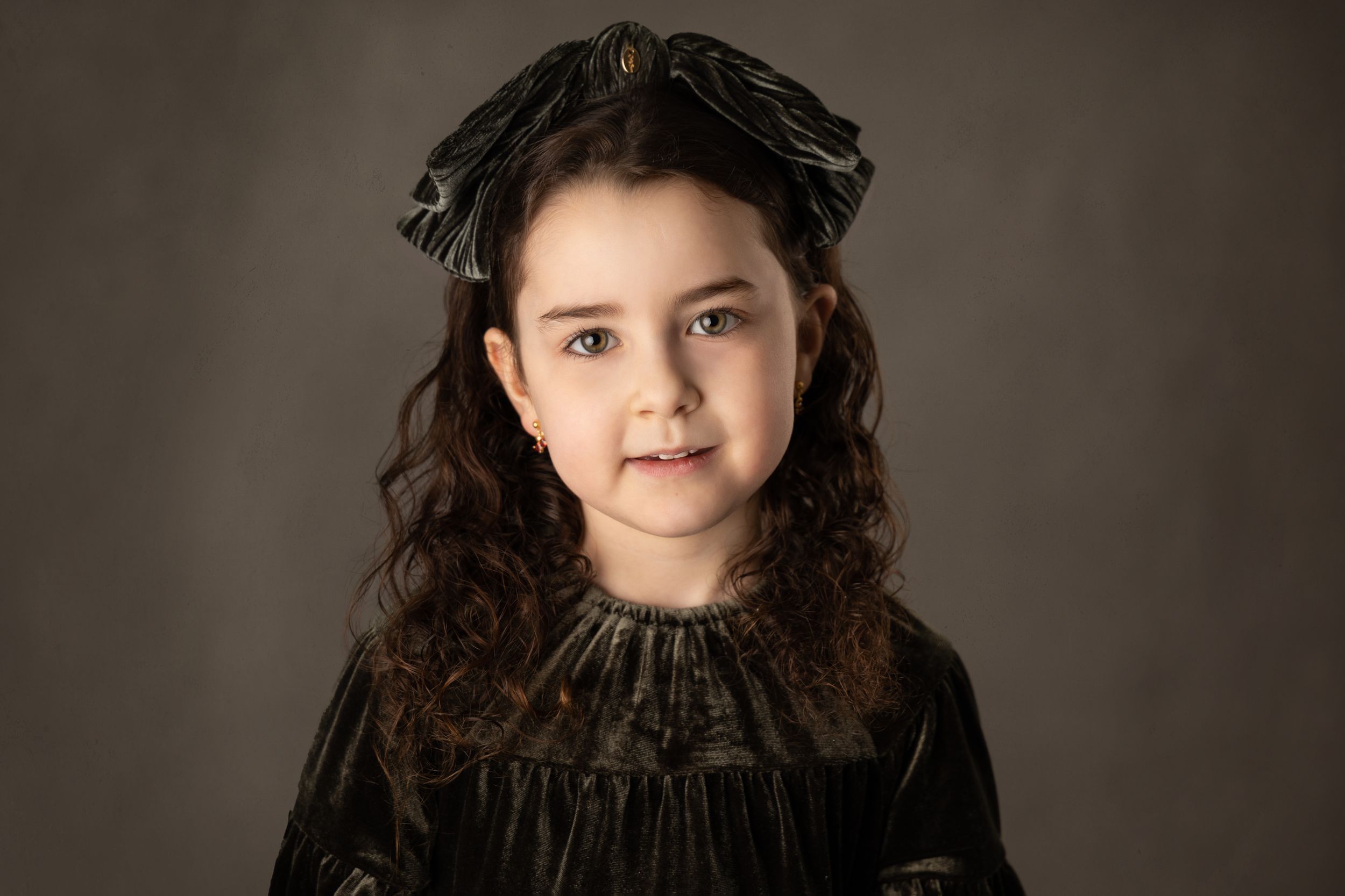 Classic portrait in black velvet dress with hair bow against dark backdrop.