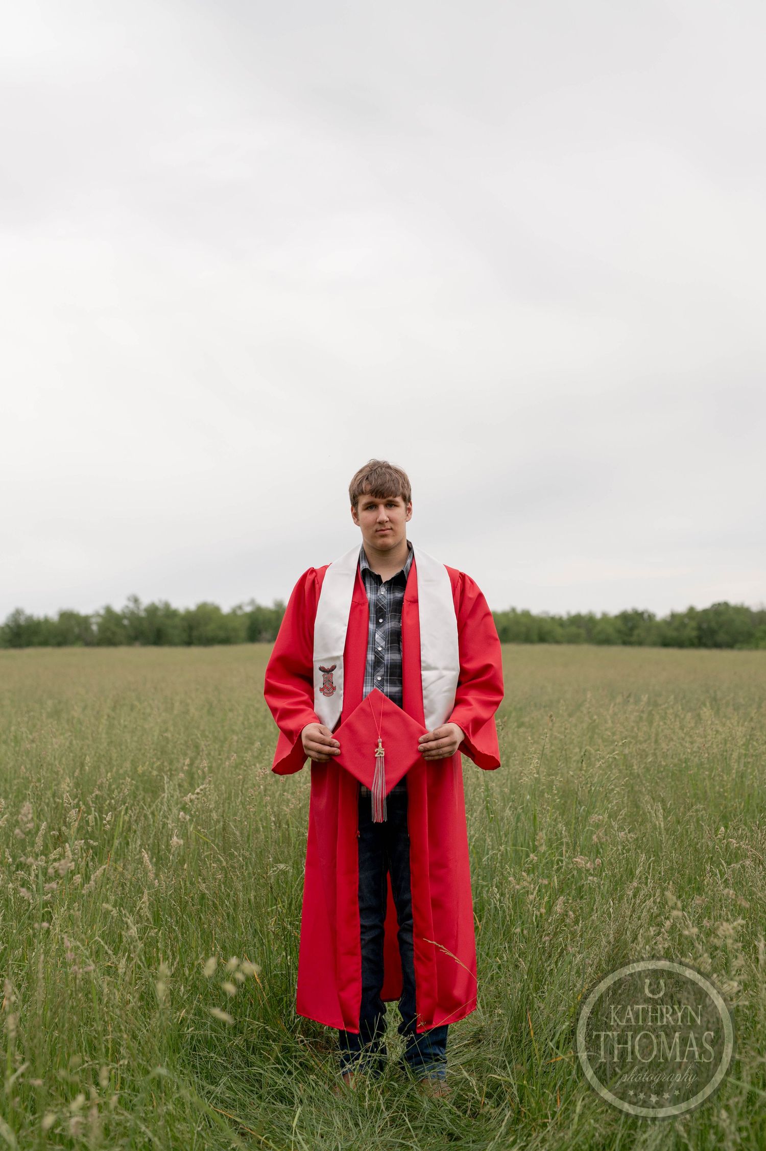 Graduate in red cap and gown stands in a green meadow under overcast sky during outdoor graduation photo session.