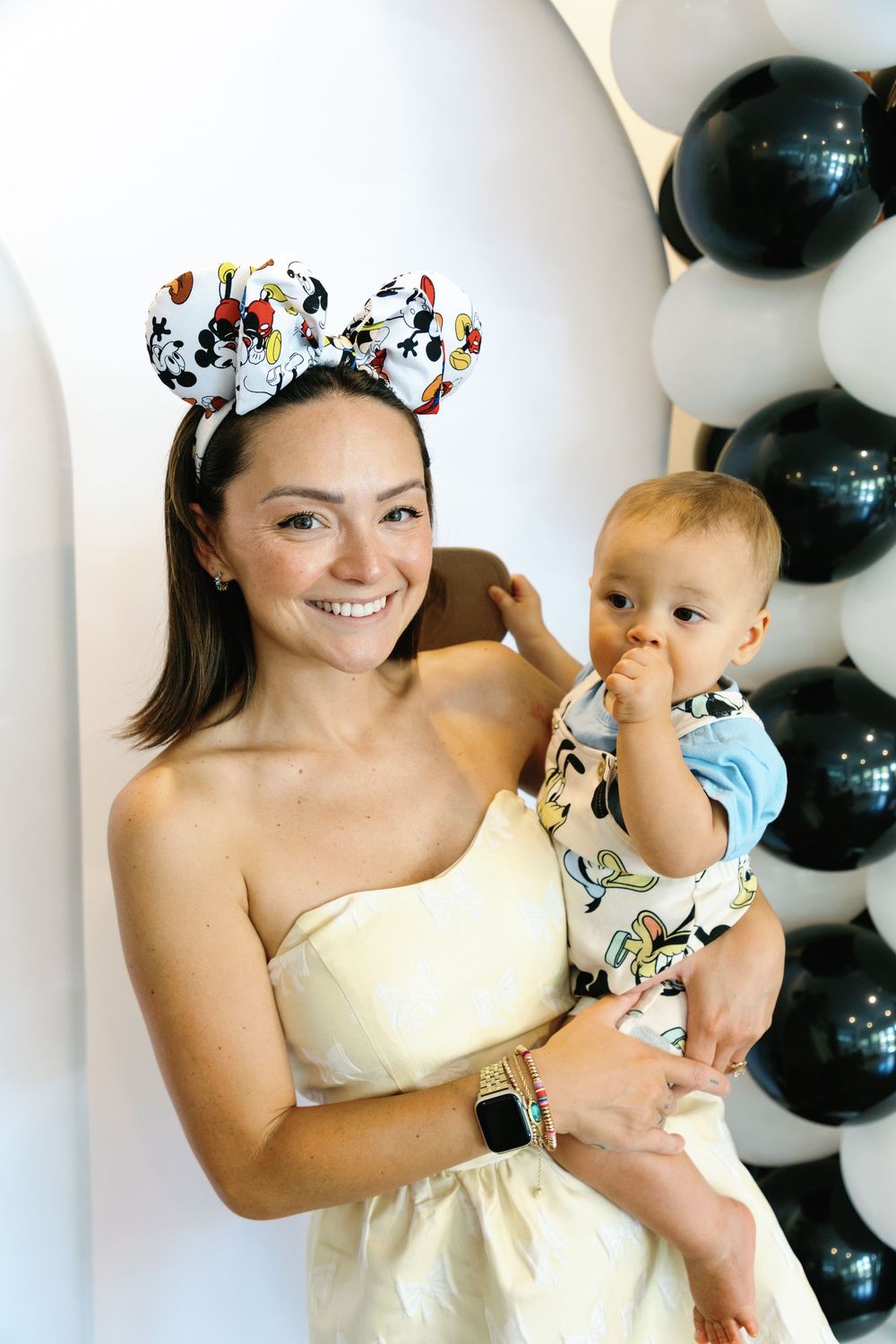 Smiling mother wearing Mickey Mouse ears holds baby at Disney-themed first birthday party.
