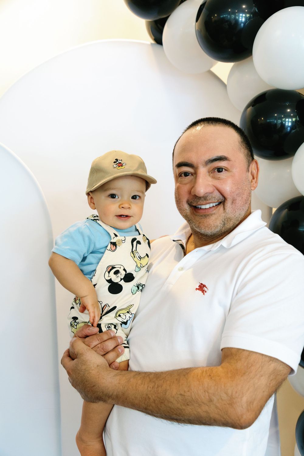 Smiling father holds baby dressed in Mickey Mouse outfit at first birthday party.
