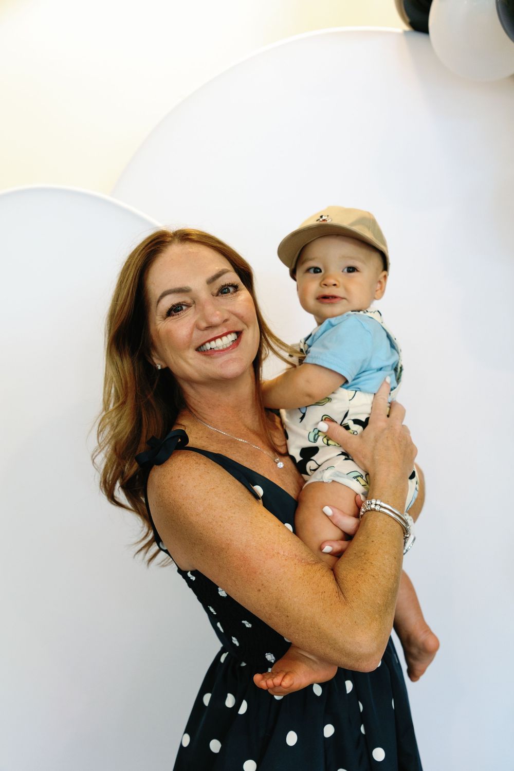 Smiling woman in polka dot dress holding an adorable baby boy in Mickey Mouse outfit at a Disney themed birthday party.