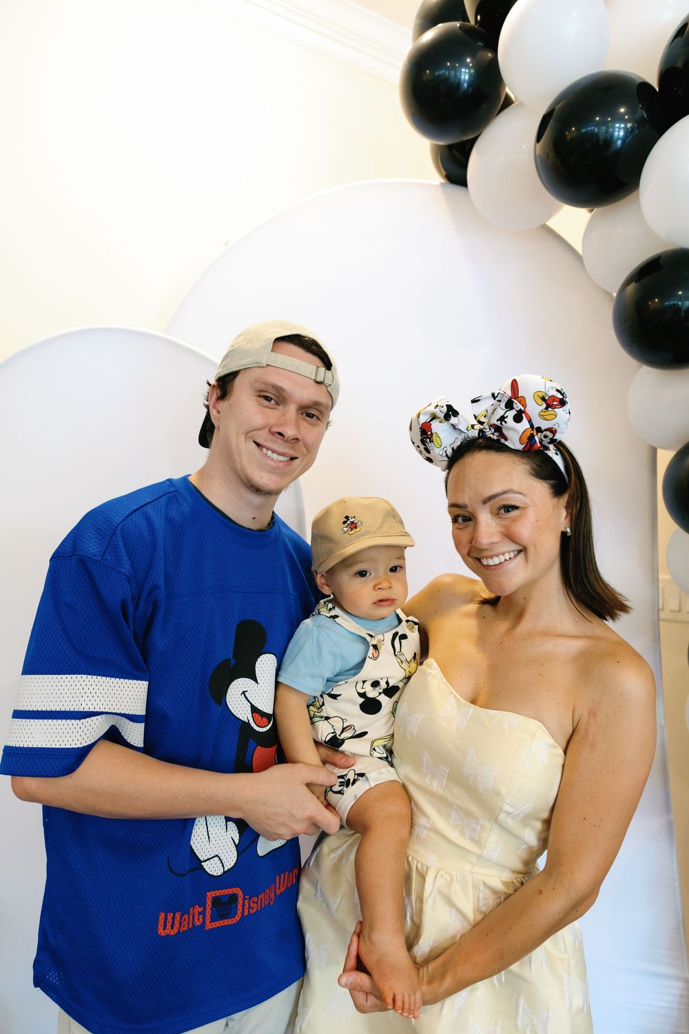 Happy couple holding baby boy in Mickey Mouse romper posing in front of black and white balloon arch at first birthday party.