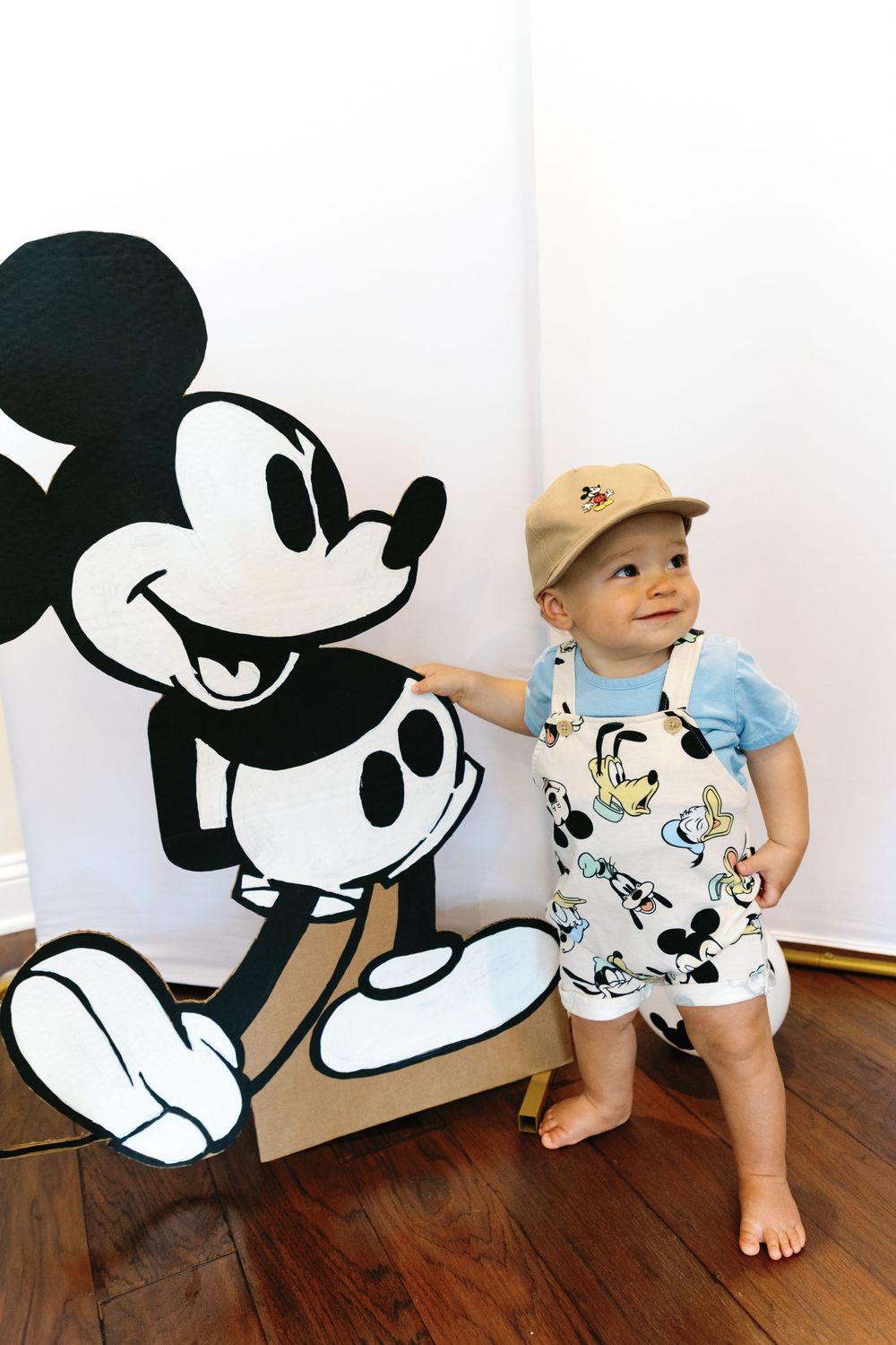 Adorable baby in Mickey Mouse overalls stands beside large Mickey Mouse cardboard cutout.